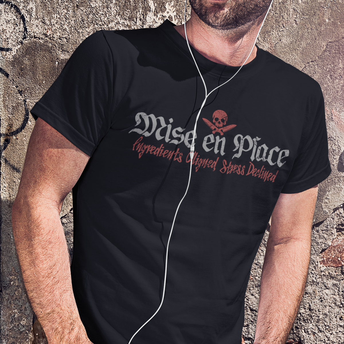 A man wearing a black short-sleeve graphic T-shirt with the words 'Miser en Place' and a skull motif in white and red, paired with blue jeans, standing against a textured stone wall.