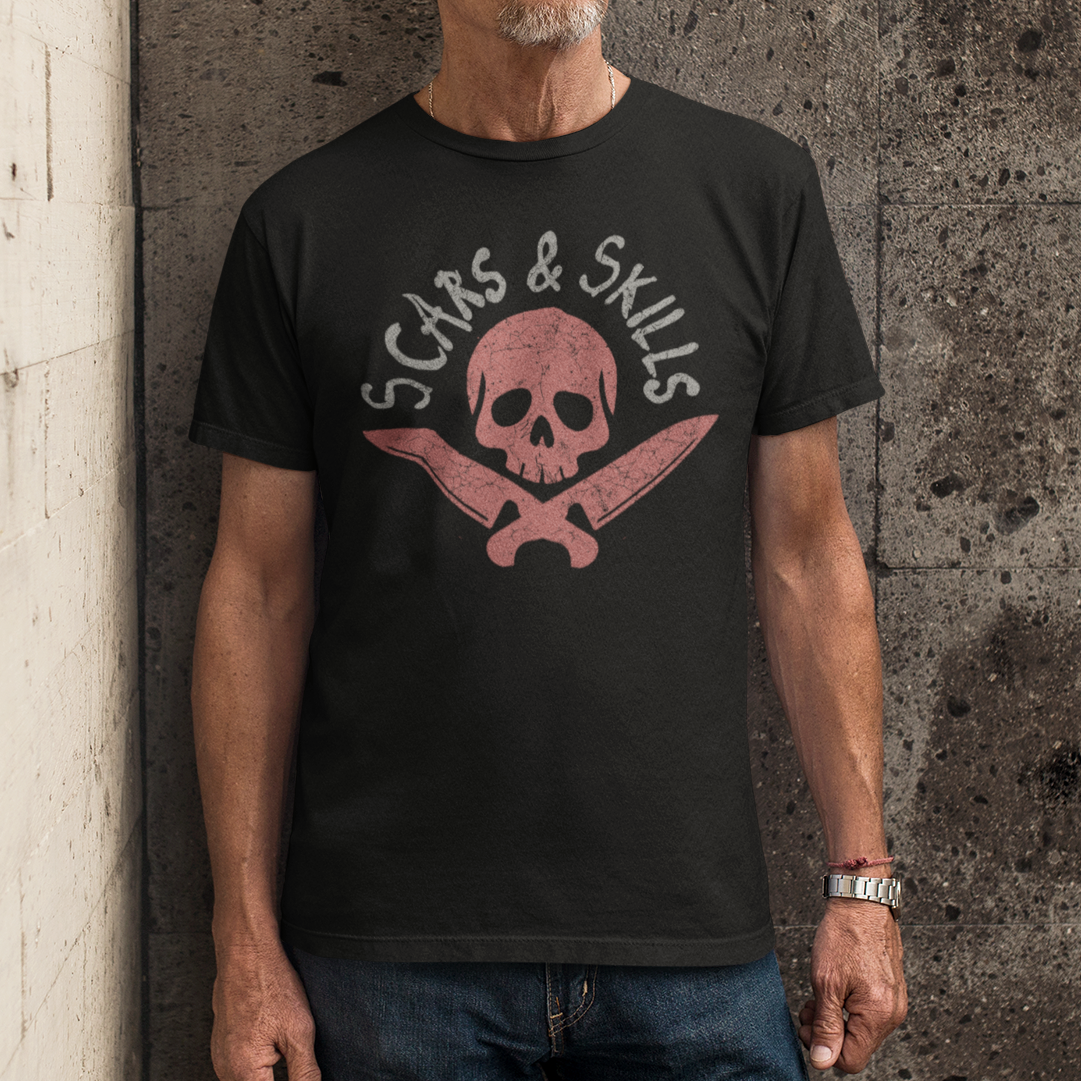 A man wearing a black graphic t-shirt with a pink skull and crossbones design and the text 'SCARS & SKILLS' stands against a textured concrete wall.