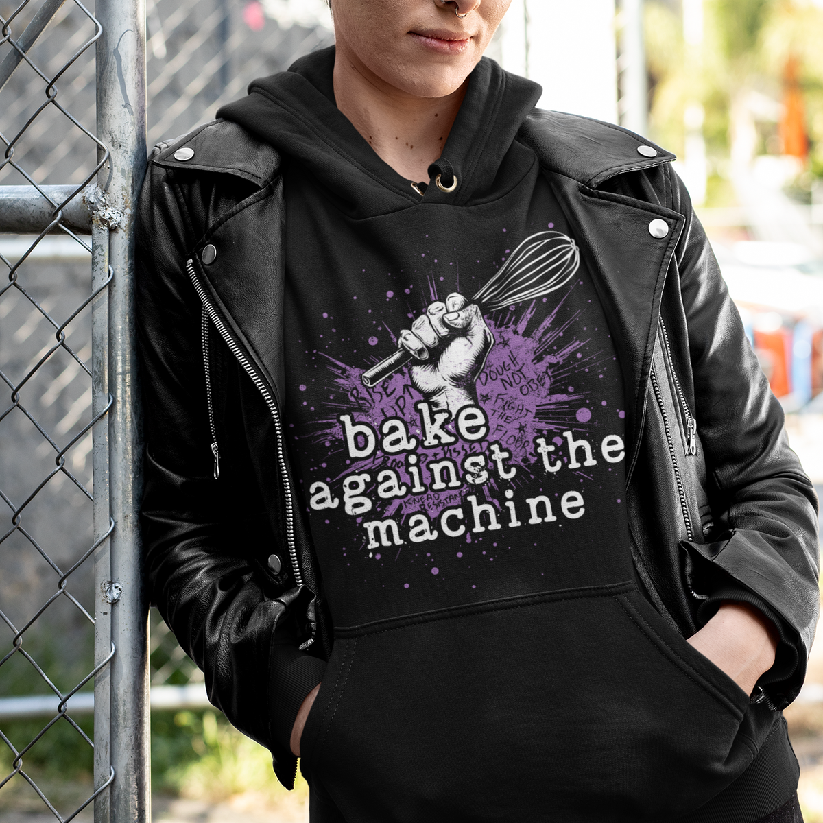A person wearing a black hoodie with a purple and white graphic print that reads 'bake against the machine' and a black leather jacket, standing outdoors next to a chain-link fence.