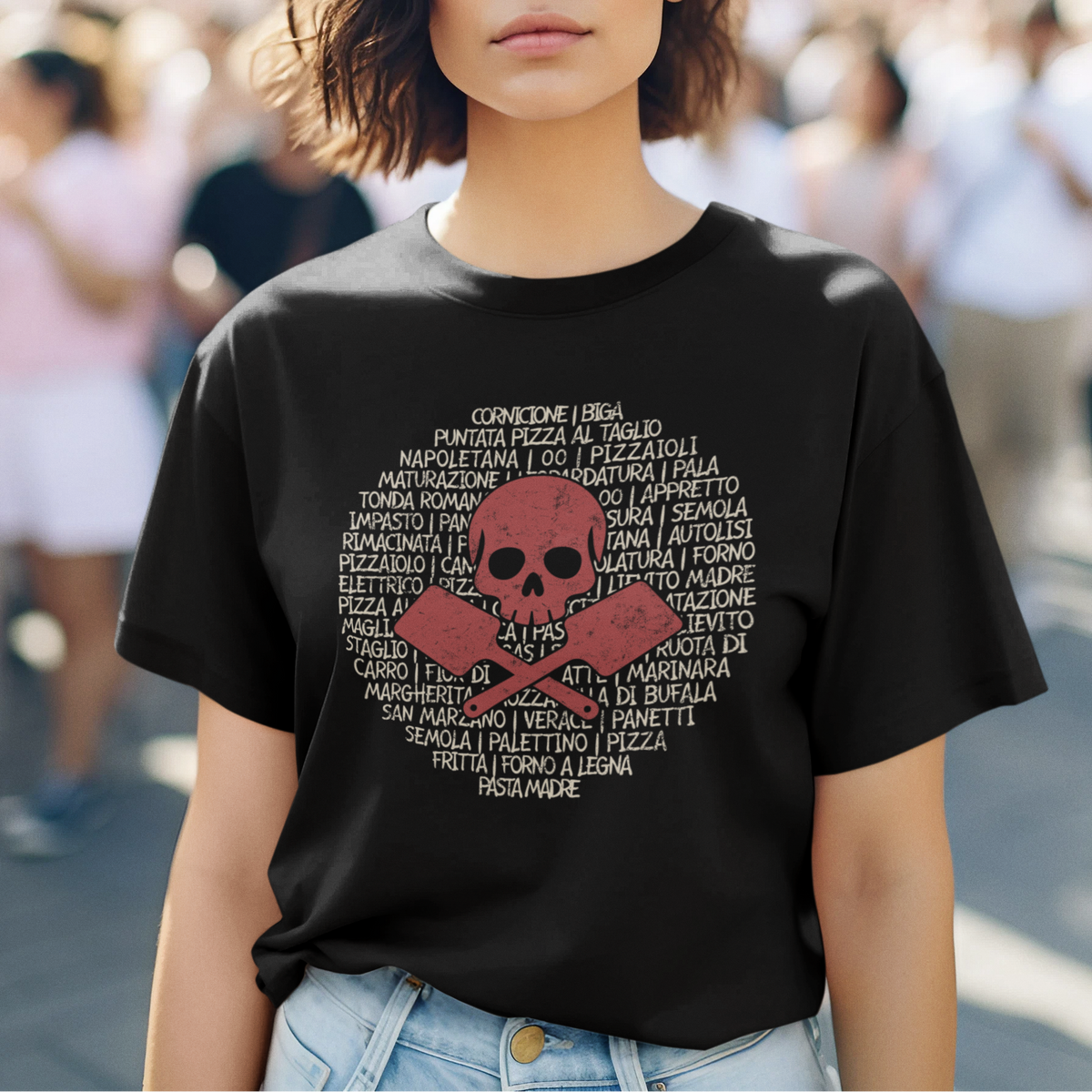 A woman wearing a black graphic T-shirt with a red skull and crossbones design and white text, photographed outdoors with a blurred crowd and building in the background.