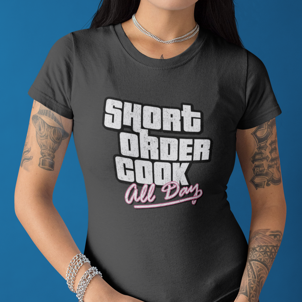A woman wearing a dark gray graphic t-shirt with the text 'SHORT ORDER COOK All Day' in white and pink, posed against a solid blue background.