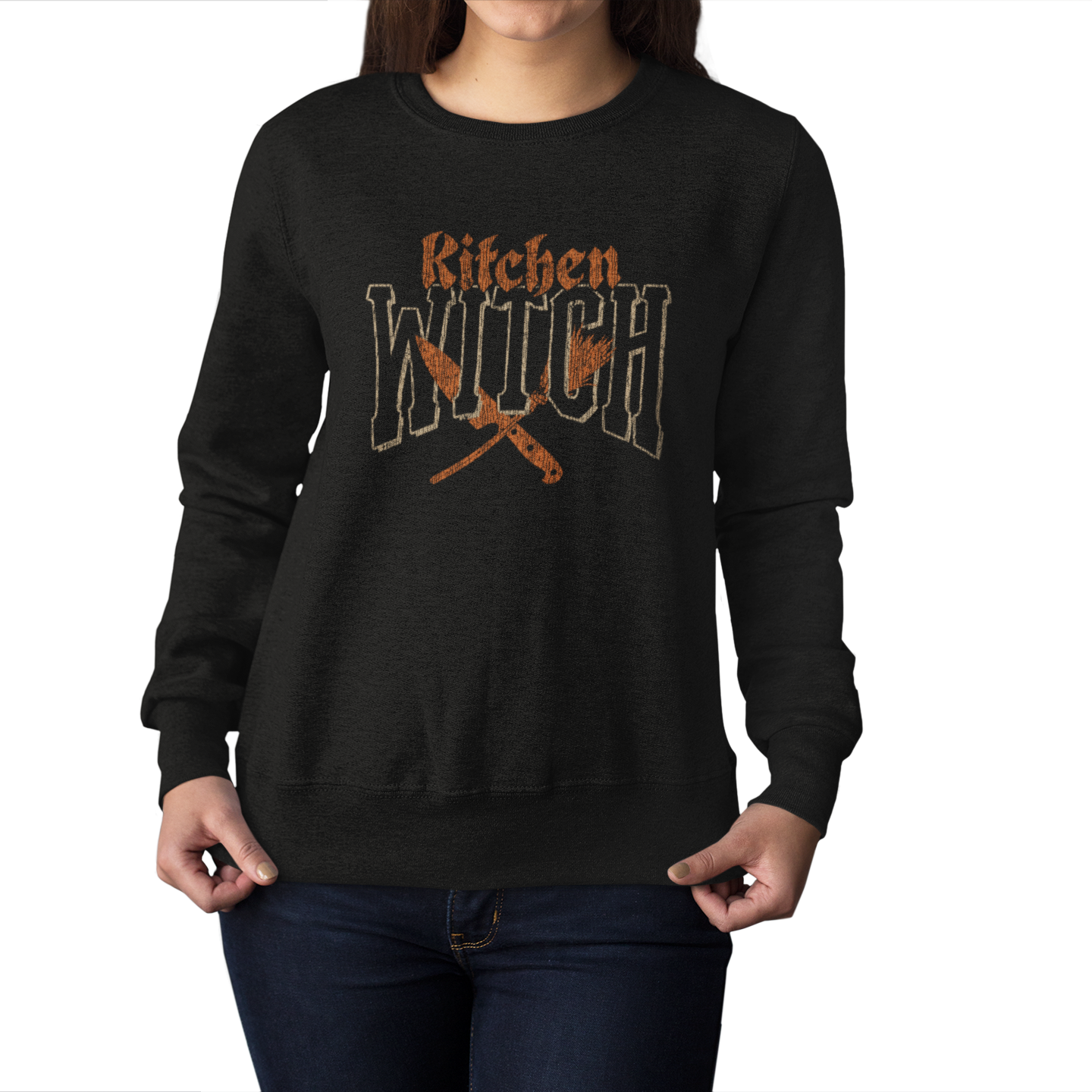 A woman wearing a black crewneck sweatshirt with a distressed orange graphic that reads 'Kitchen Witch' above crossed kitchen utensils, shown against a plain white background.