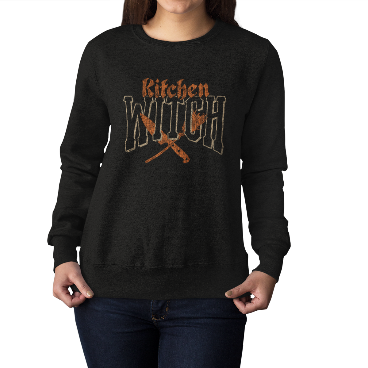 A woman wearing a black crewneck sweatshirt with a distressed orange graphic that reads 'Kitchen Witch' above crossed kitchen utensils, shown against a plain white background.