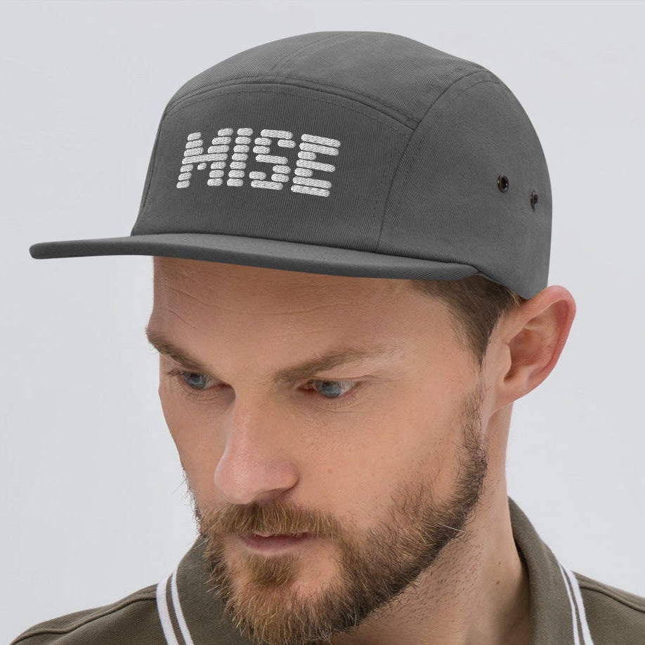 Man wearing a brown polo shirt and gray cap with 'MISE' logo on a light gray background