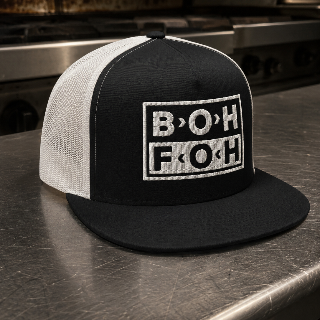 A black and white trucker hat with a black front panel and white mesh back, featuring a white embroidered graphic on the front that reads 'B>OHFOH'. The hat is placed on a stainless steel counter in a commercial kitchen setting.