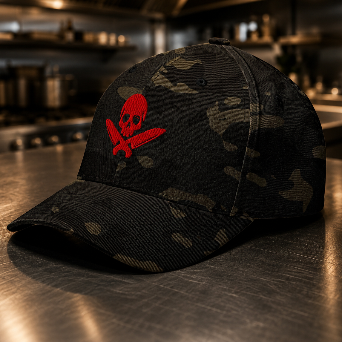 A black camouflage baseball cap with a red skull and crossed knives logo on the front, resting on a metallic kitchen counter with a blurred professional kitchen in the background.