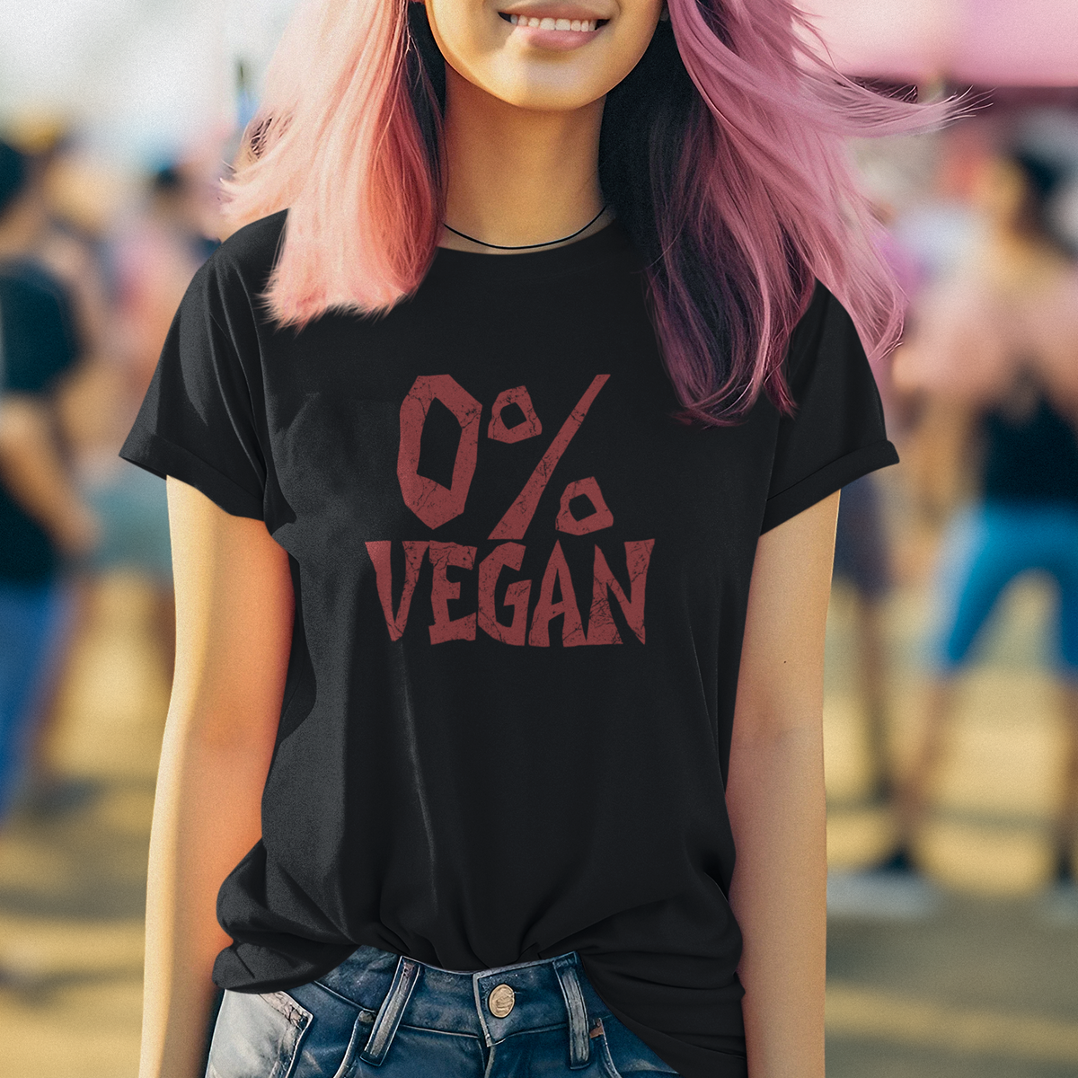 A smiling woman with pink and dark purple hair stands outdoors wearing a black graphic T-shirt that reads '0% VEGAN' in red, paired with blue jeans. The background is a softly blurred outdoor crowd scene.