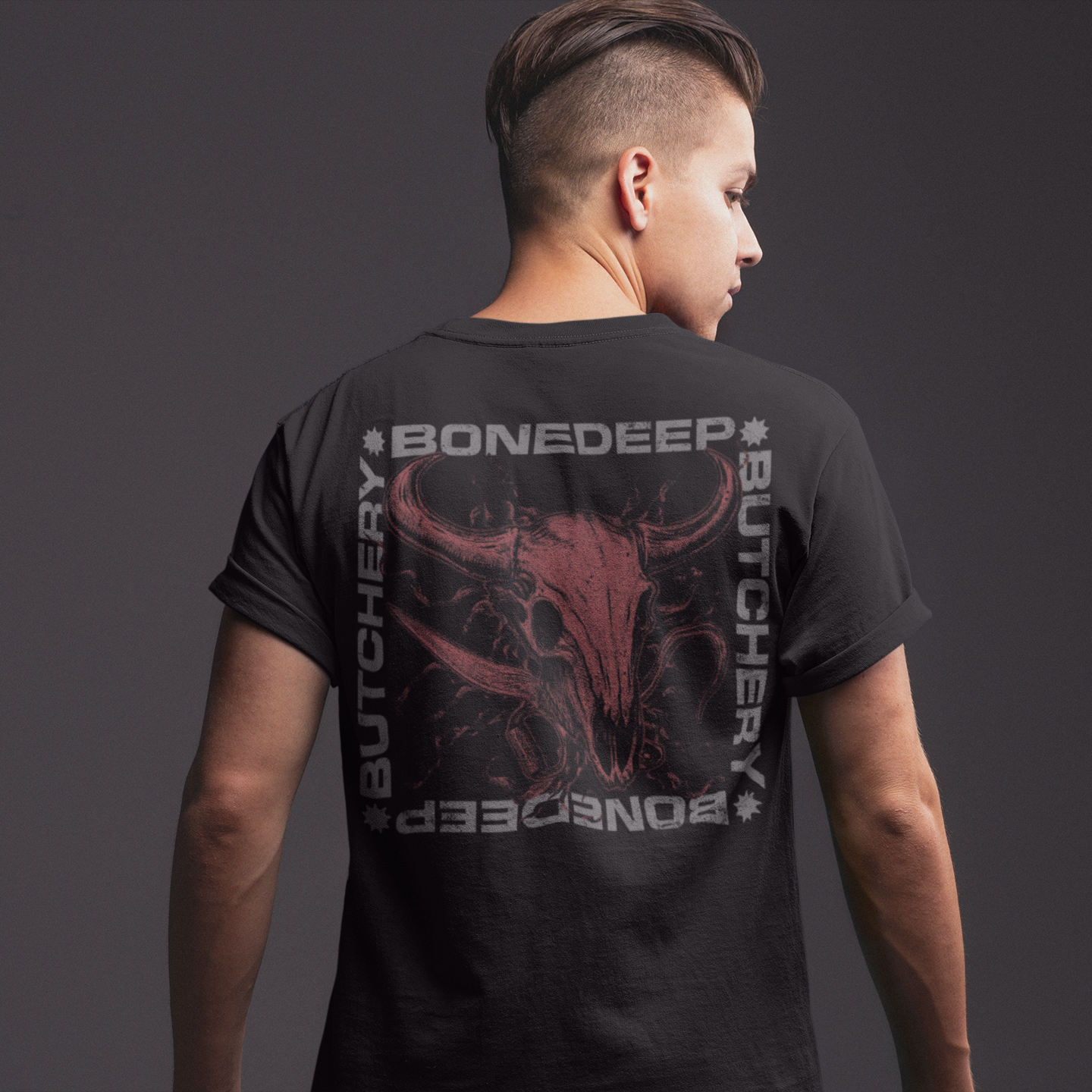 A man shown from the back wearing a black t-shirt with a large graphic print on the back. The graphic features a red bull skull in the center, surrounded by the text 'BONEDEEP' and 'BUTCHERY' in a distressed gray font, set against a dark gray studio background.
