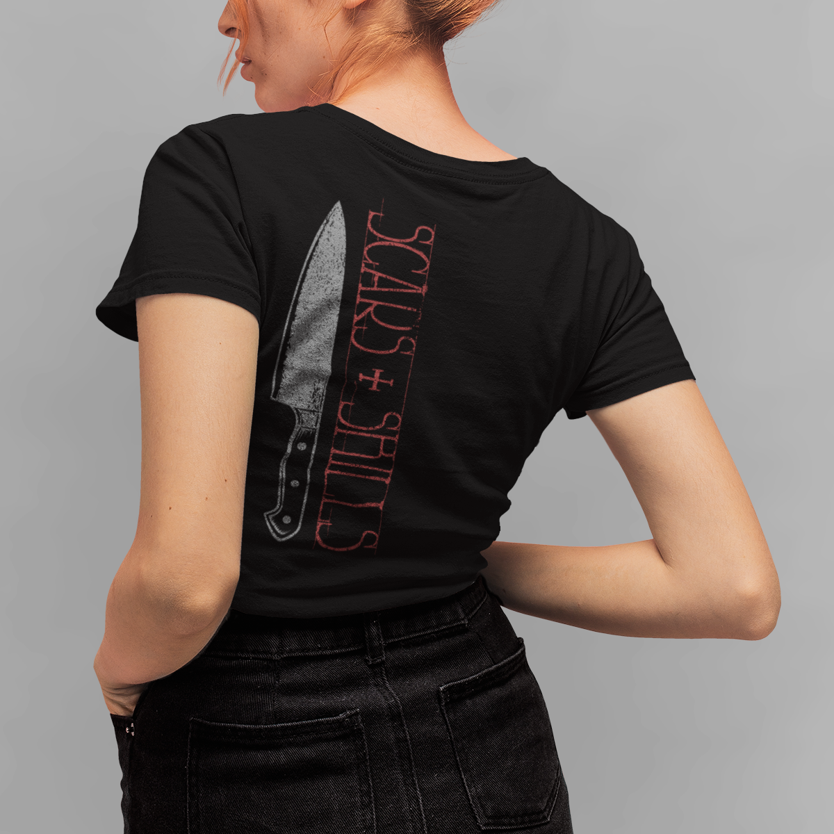 A woman with orange hair in a bun is shown from the back wearing a black t-shirt with a graphic of a knife and the text 'SCARS + SHINERS' in red, paired with black jeans, against a solid gray background.