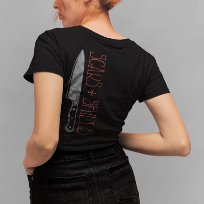 A woman with orange hair in a bun is shown from the back wearing a black t-shirt with a graphic of a knife and the text 'SCARS + SHINERS' in red, paired with black jeans, against a solid gray background.