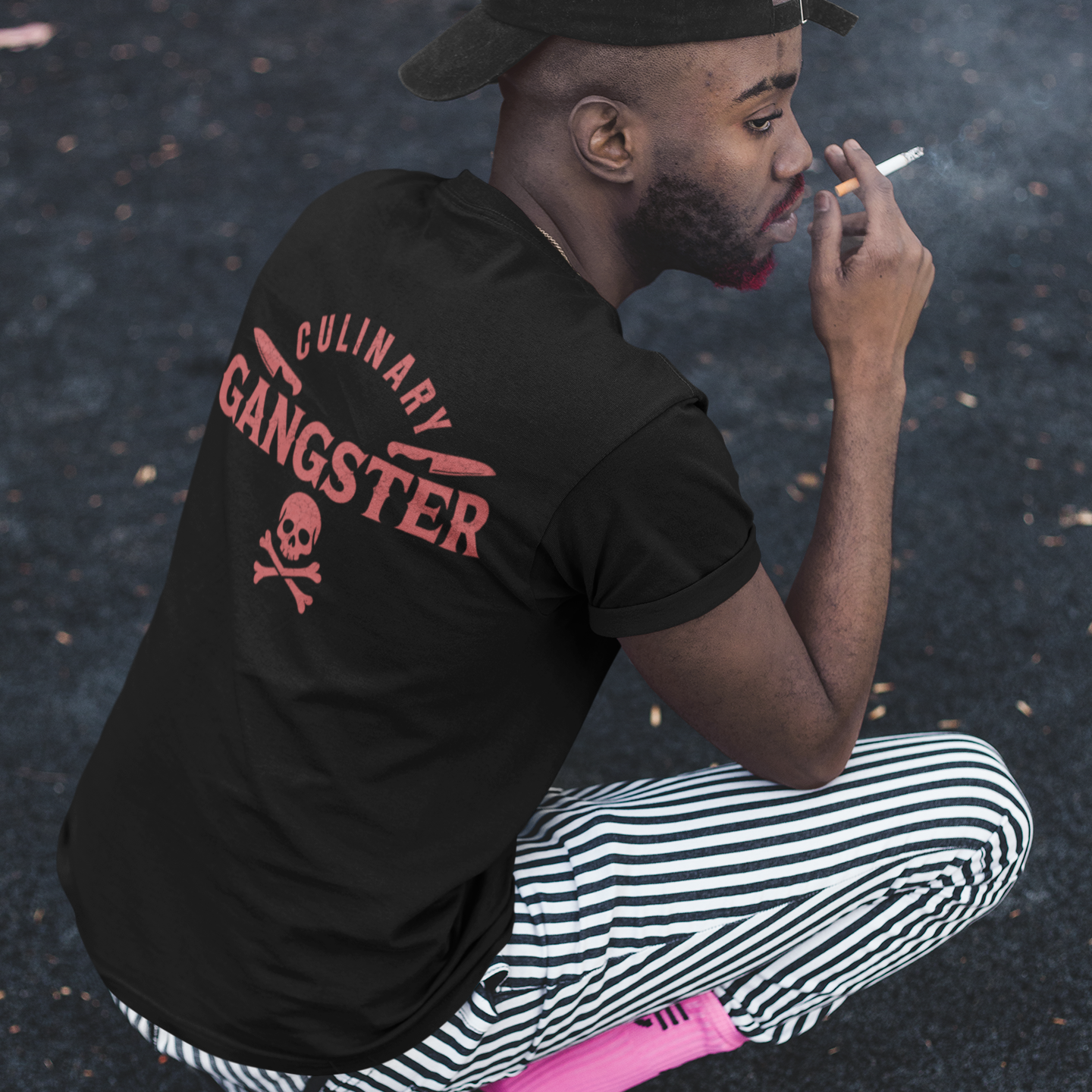 A man crouching outdoors on asphalt, wearing a black t-shirt with a red 'Culinary Gangster' graphic on the back, black and white striped pants, and a black cap.
