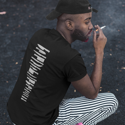 A man crouching on asphalt wearing a black t-shirt with a white vertical graphic print on the back, black-and-white striped pants, a black cap, and black sneakers, while smoking a cigarette.