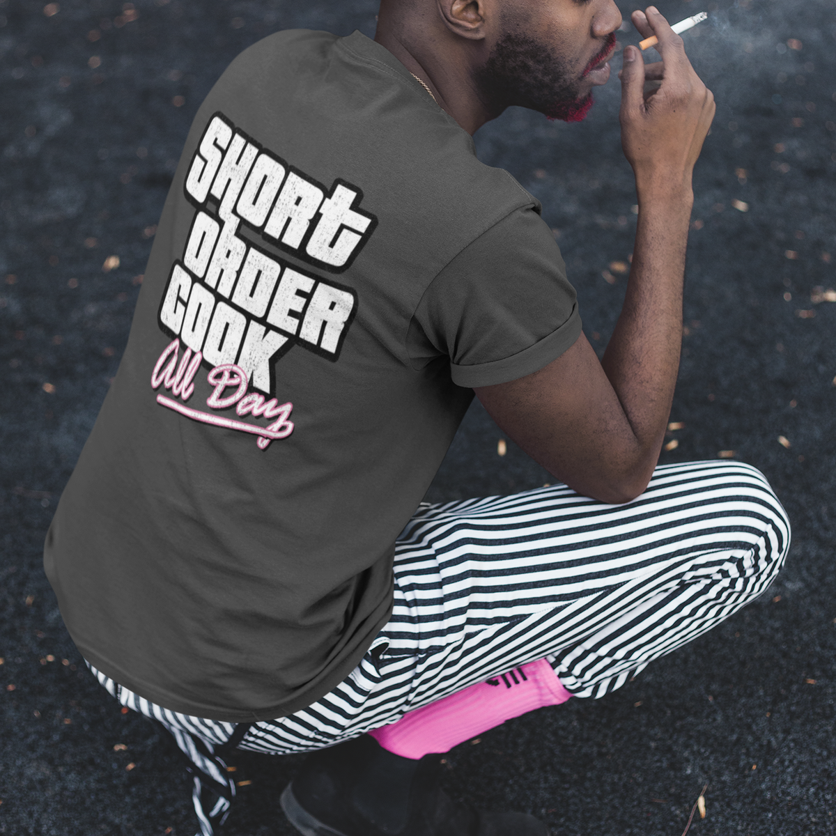 A man crouching outdoors on asphalt, wearing a dark gray t-shirt with 'SHORT ORDER COOK All Day' printed in white and pink on the back, paired with black and white striped pants and a black cap.