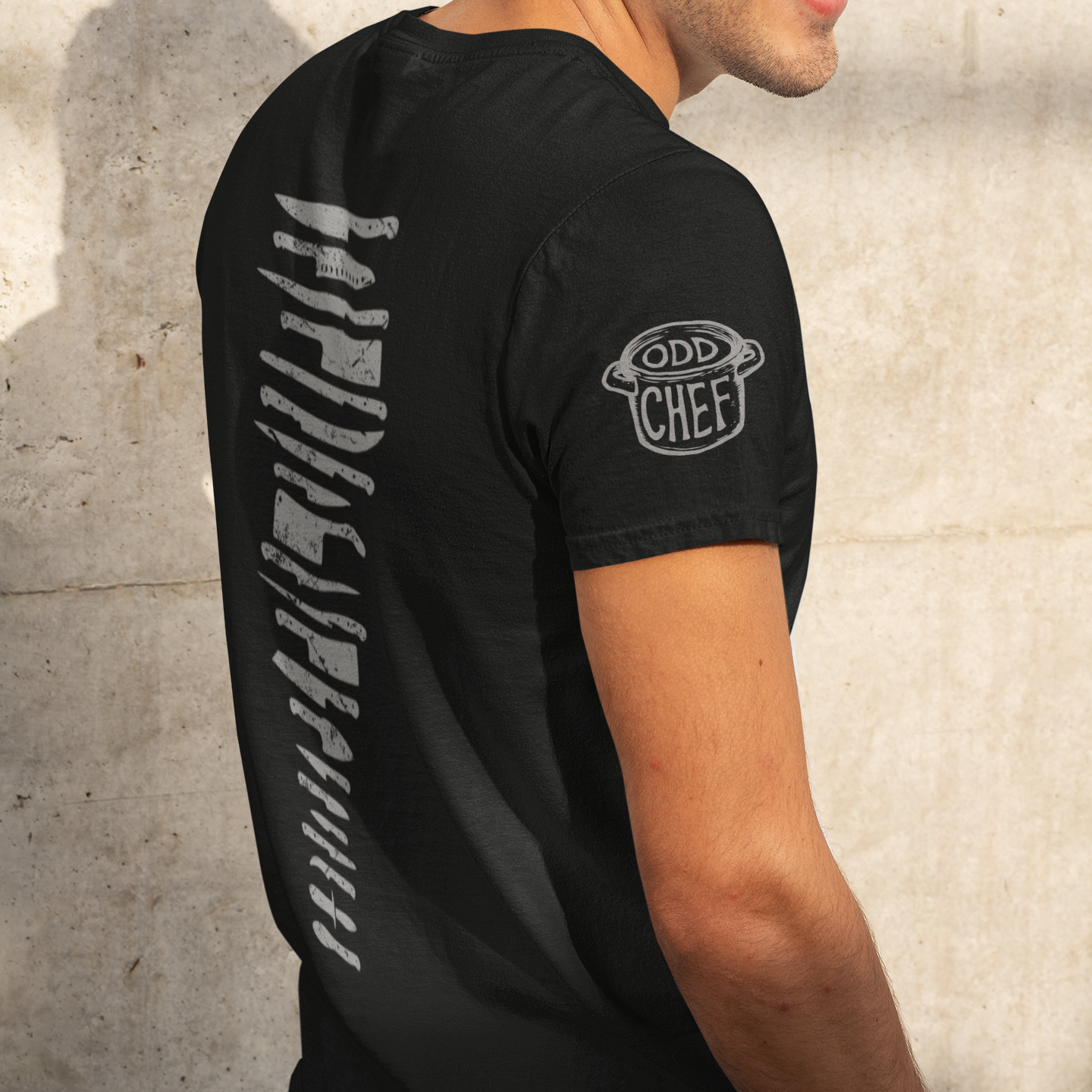 A man wearing a black t-shirt with a vertical white graphic on the back and a 'Odd Chef' logo on the left sleeve, standing against a textured concrete wall.