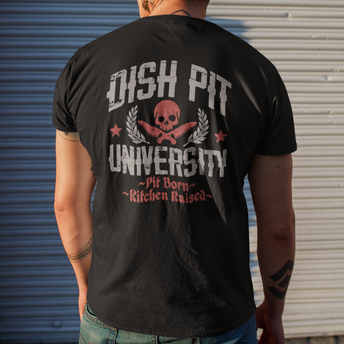 A man viewed from behind wearing a black graphic t-shirt with a distressed white and red 'OHSH PIT UNIVERSITY' design on the back, standing in front of a corrugated metal garage door.