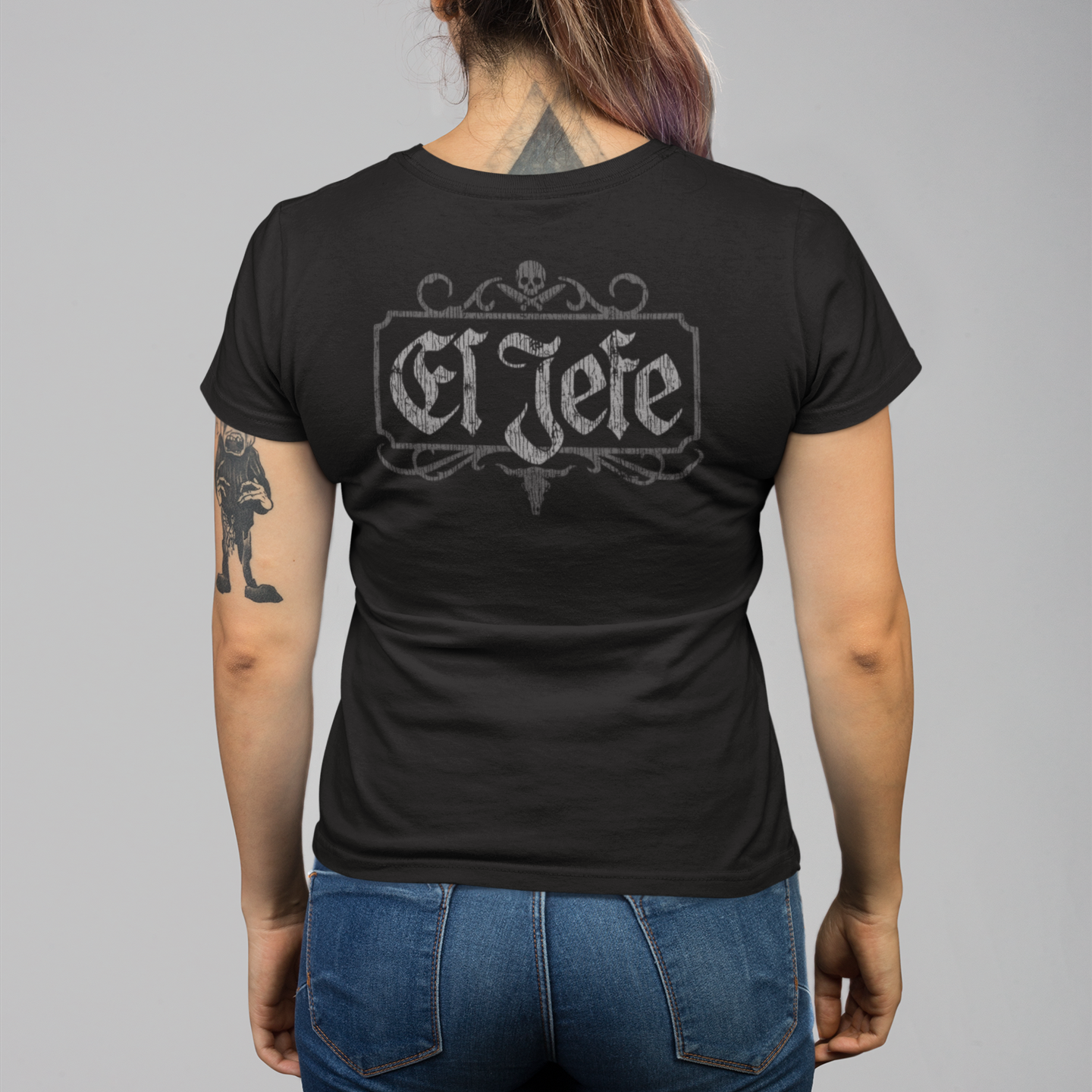 A woman viewed from the back wearing a black short-sleeve t-shirt with a gray 'El Jefe' gothic-style graphic on the back, paired with blue jeans, against a solid gray studio background.
