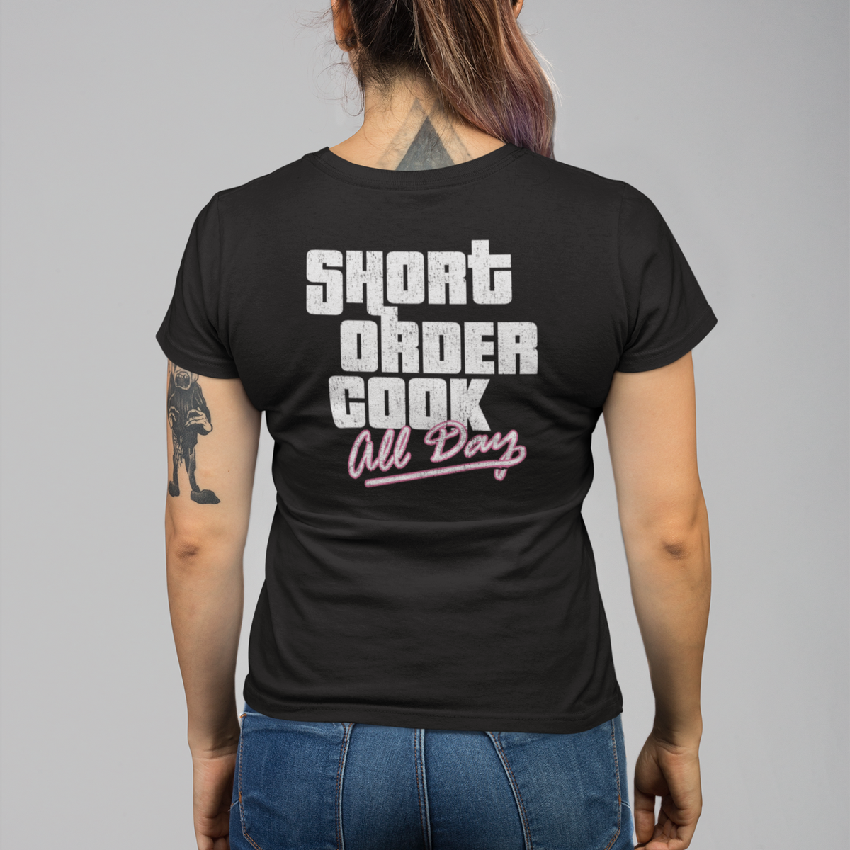 A woman viewed from behind wearing a black t-shirt with a graphic that reads 'SHORT ORDER COOK All Day' in white and pink, paired with blue jeans, against a solid gray background.