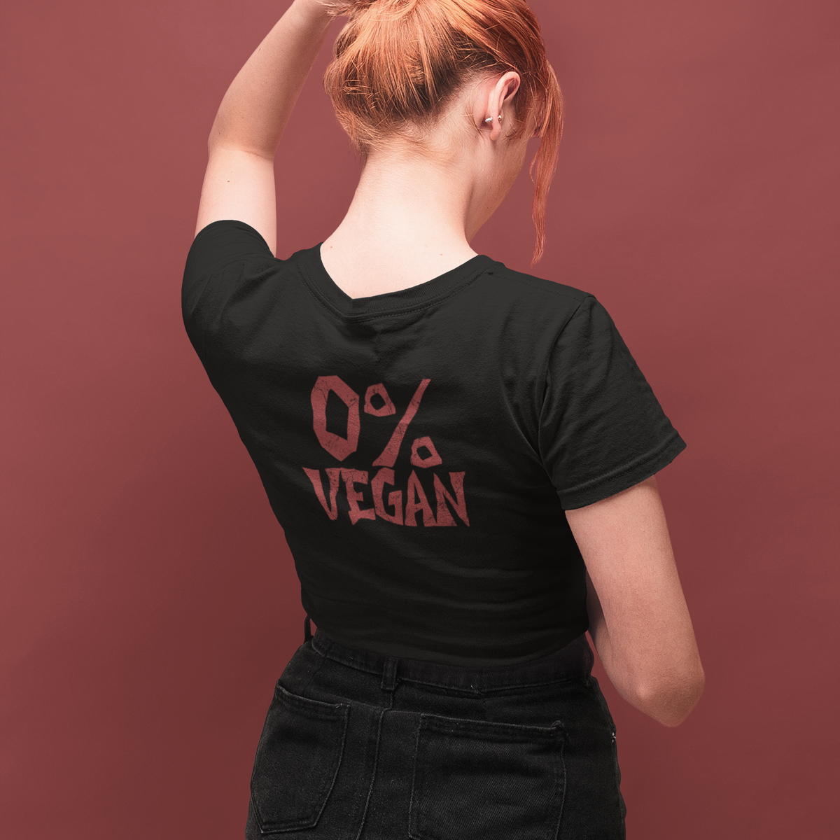 A woman with red hair in a bun is shown from behind wearing a black t-shirt with a red graphic that reads '0% VEGAN'. She is posing against a solid dark reddish-brown background.
