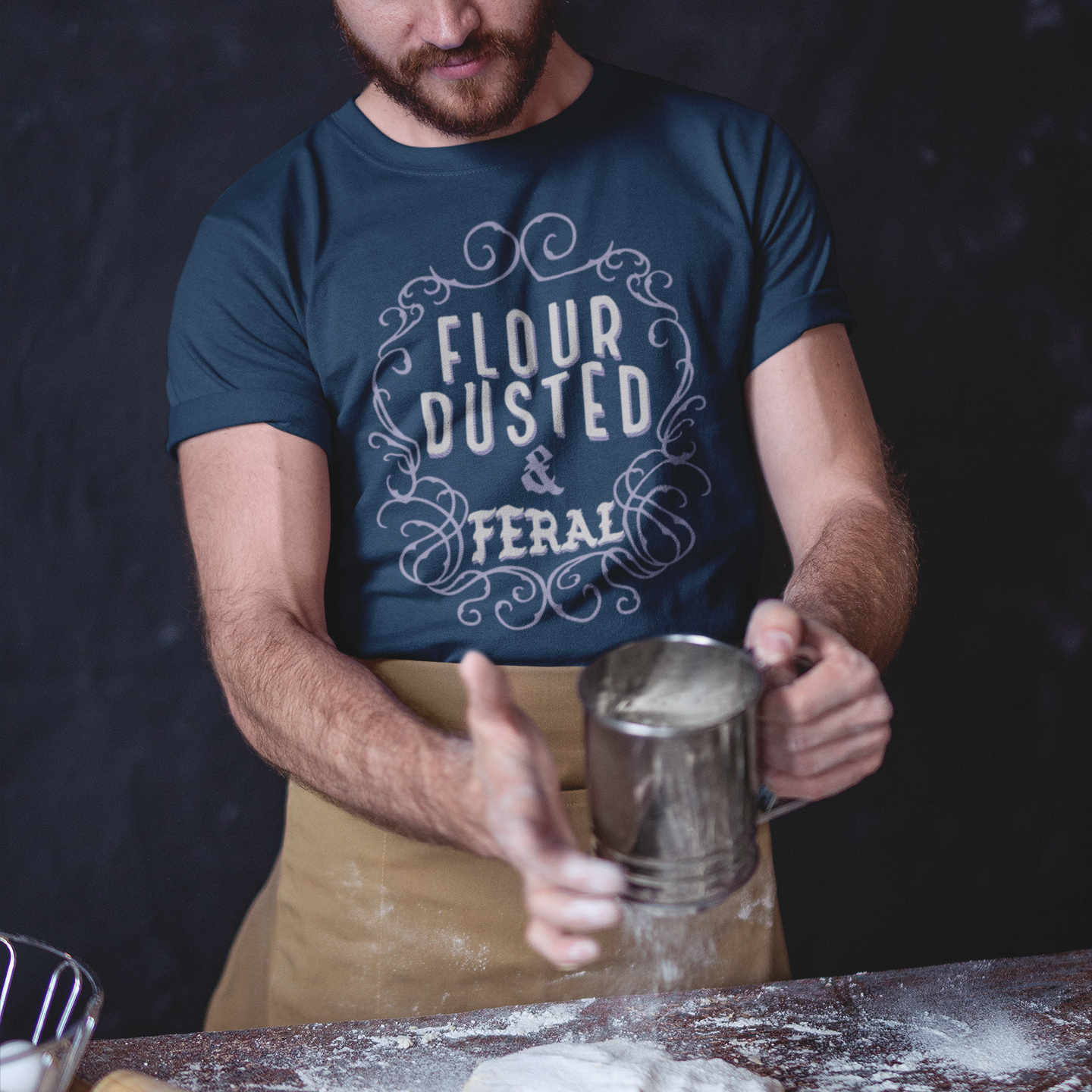Man in a kitchen wearing a 'Flour Dusted & Feral' shirt, preparing dough.