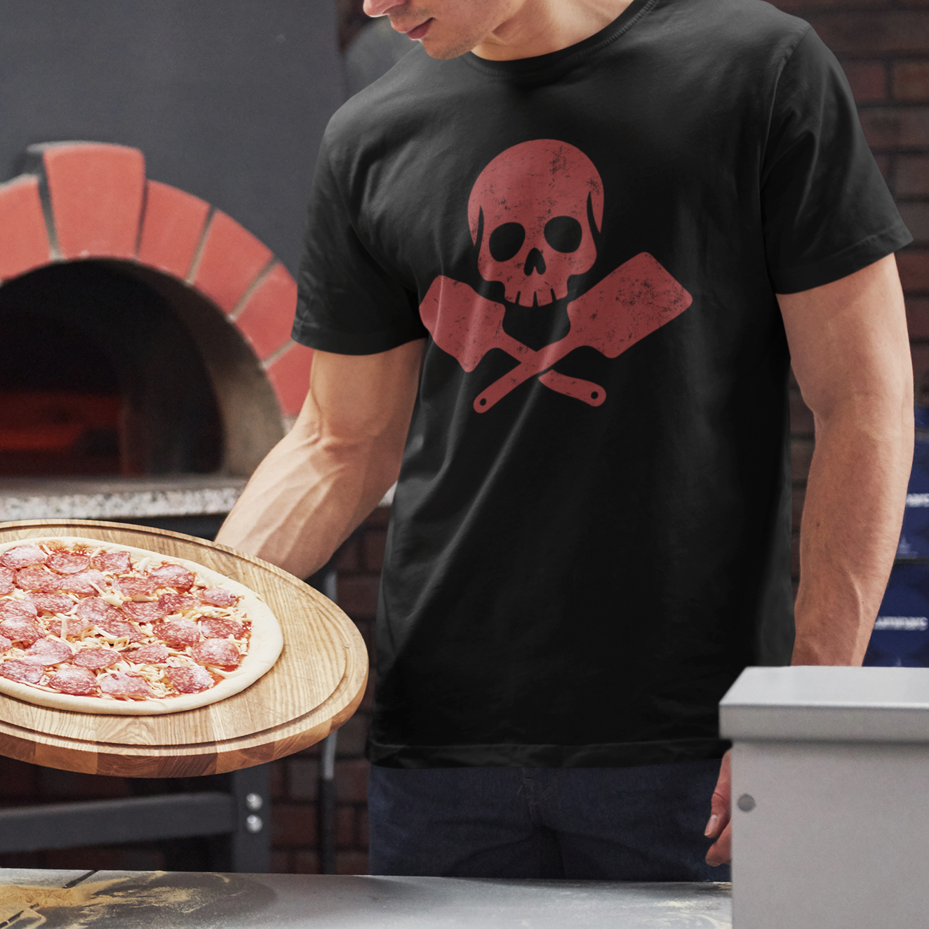 A man in a black graphic t-shirt holding a freshly baked pizza on a wooden board in front of a brick pizza oven.