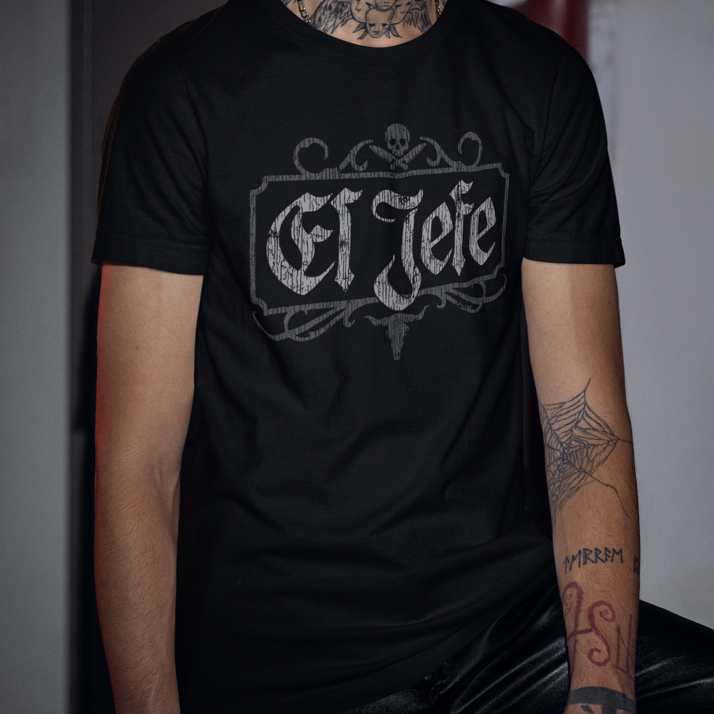 A man wearing a black short-sleeve t-shirt with a distressed gray 'El Jefe' gothic-style graphic on the chest, set against a dark, moody background.