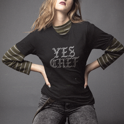 A woman kneels on a dark studio floor wearing a black graphic T-shirt with striped long sleeves and gray jeans. The shirt has the words 'YES CHEF' printed across the chest.