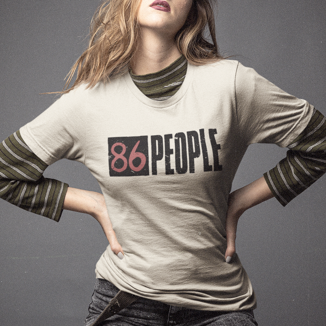 A woman kneels on a dark gray studio floor wearing a beige graphic t-shirt with olive green striped long sleeves and dark gray jeans. The shirt features the text '86 PEOPLE' in black and red block print on the chest.