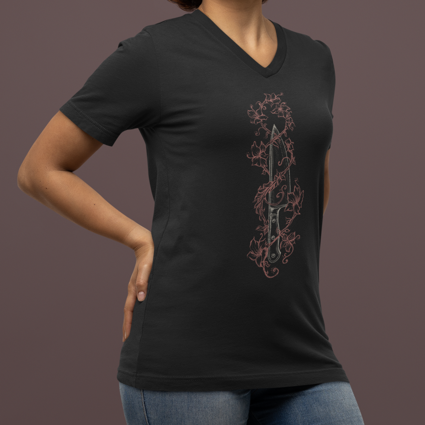 A woman wearing a black V-neck t-shirt with a vertical graphic print of a dagger surrounded by thorns, paired with blue jeans, set against a solid dark brown studio background.