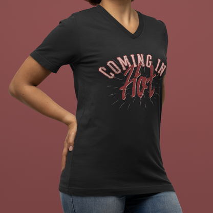 A woman wearing a black V-neck t-shirt with a red graphic that reads 'COMING IN HOT' against a solid dark reddish-brown background.