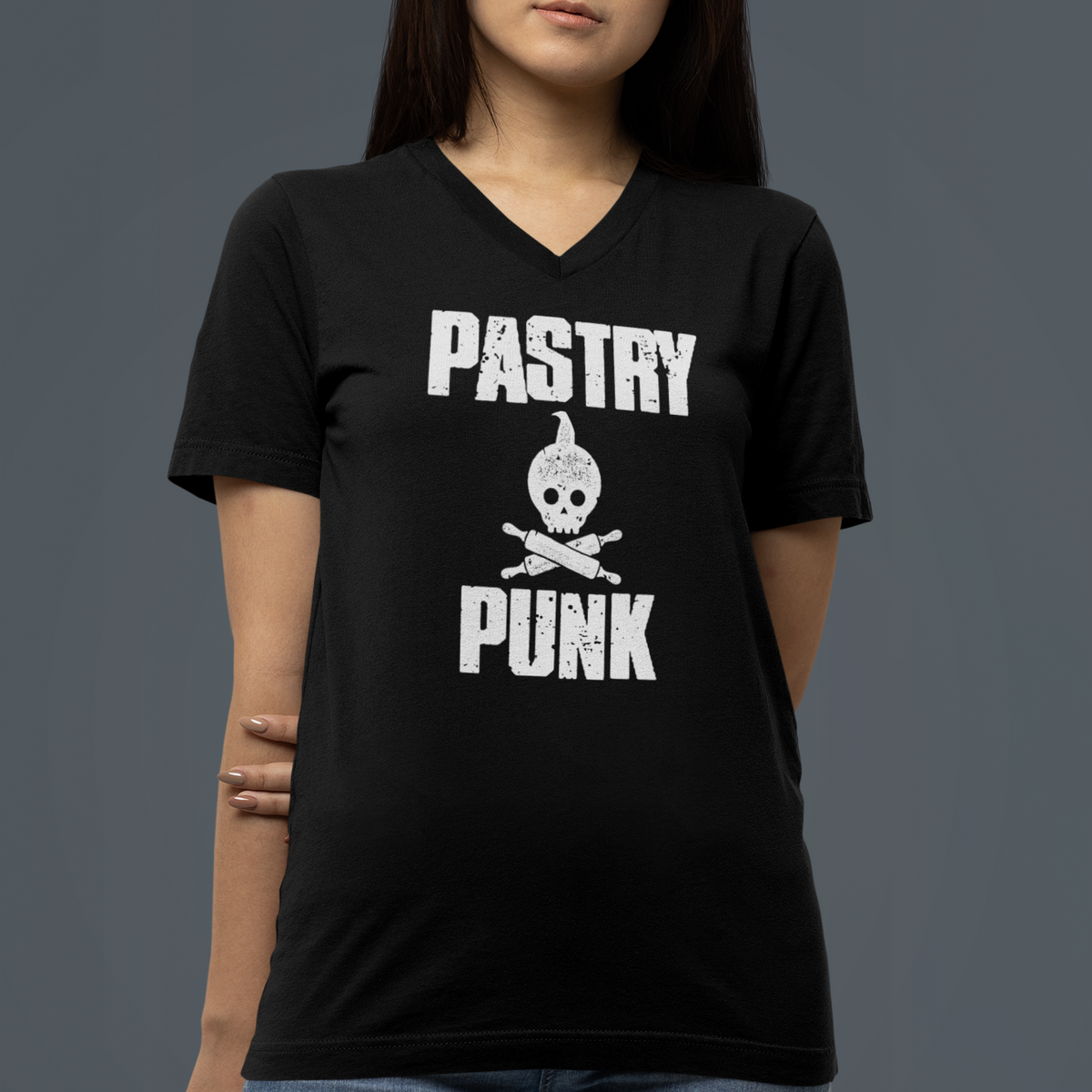 A woman wearing a black V-neck t-shirt with a white 'PASTRY PUNK' skull and crossbones graphic, paired with blue jeans, against a dark gray studio background.
