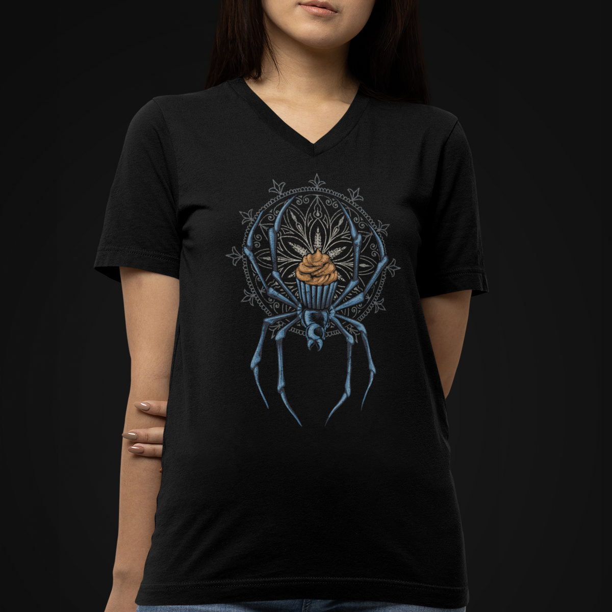 A woman wearing a black V-neck t-shirt with a detailed graphic of a blue spider holding a cupcake, paired with blue jeans, set against a solid black background.