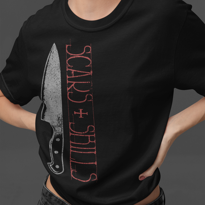 A woman wearing a black graphic t-shirt featuring a knife illustration and the text 'SCARS + SHILLS' in red, paired with dark gray jeans against a dark gray studio background.