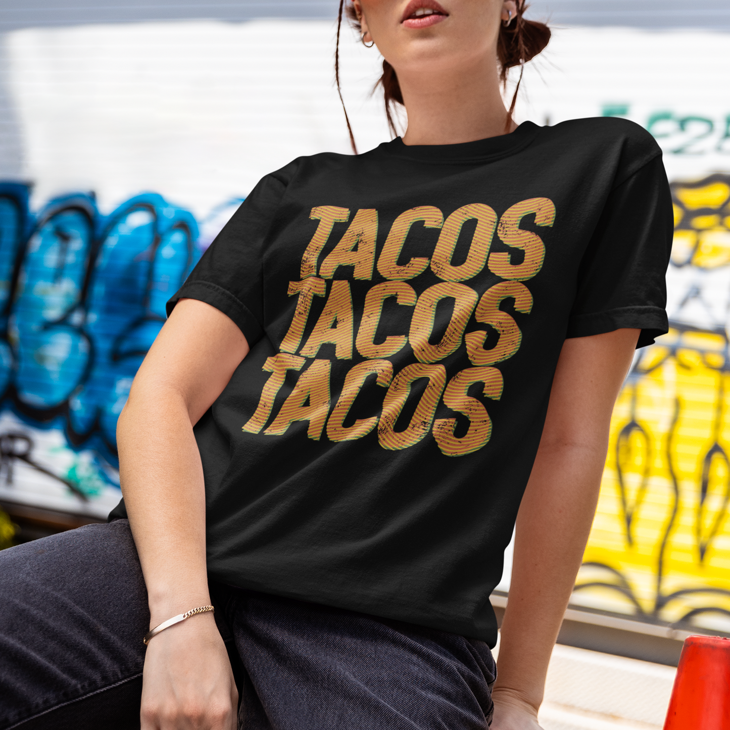 A woman wearing a black graphic t-shirt with 'TACOS TACOS TACOS' printed in a distressed gold font, posing outdoors in front of a graffiti-covered wall.