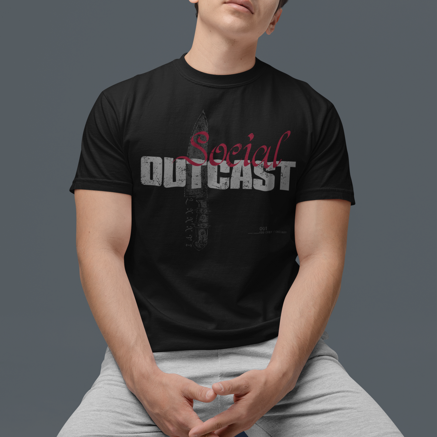 A man wearing a black graphic t-shirt with 'Social OUTCAST' printed on the front, paired with light gray shorts, posed against a solid dark gray background.