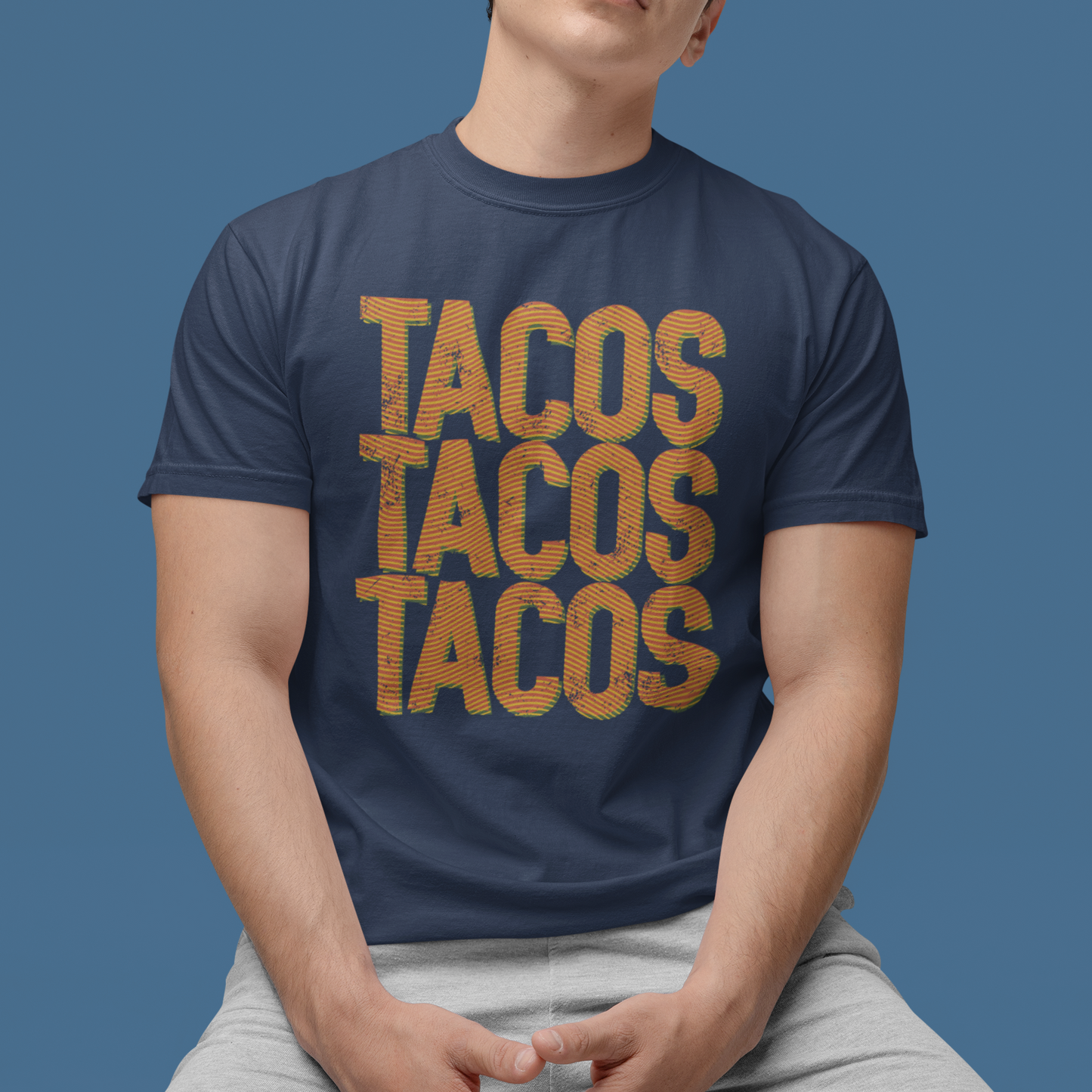 A man wearing a navy blue t-shirt with 'TACOS TACOS TACOS' printed in a distressed orange font, paired with light gray shorts, posed against a solid blue background.