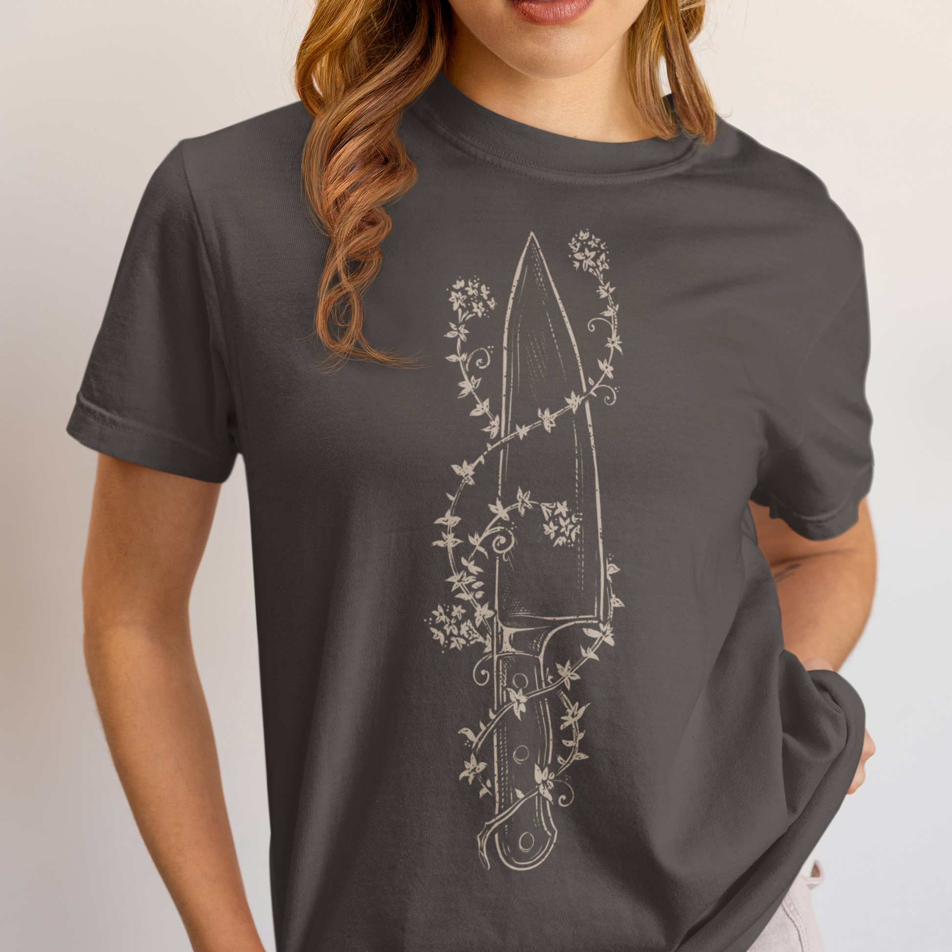 A woman wearing a dark gray t-shirt with a white line art graphic of a knife surrounded by flowers, paired with light pink jeans against a plain light gray background.
