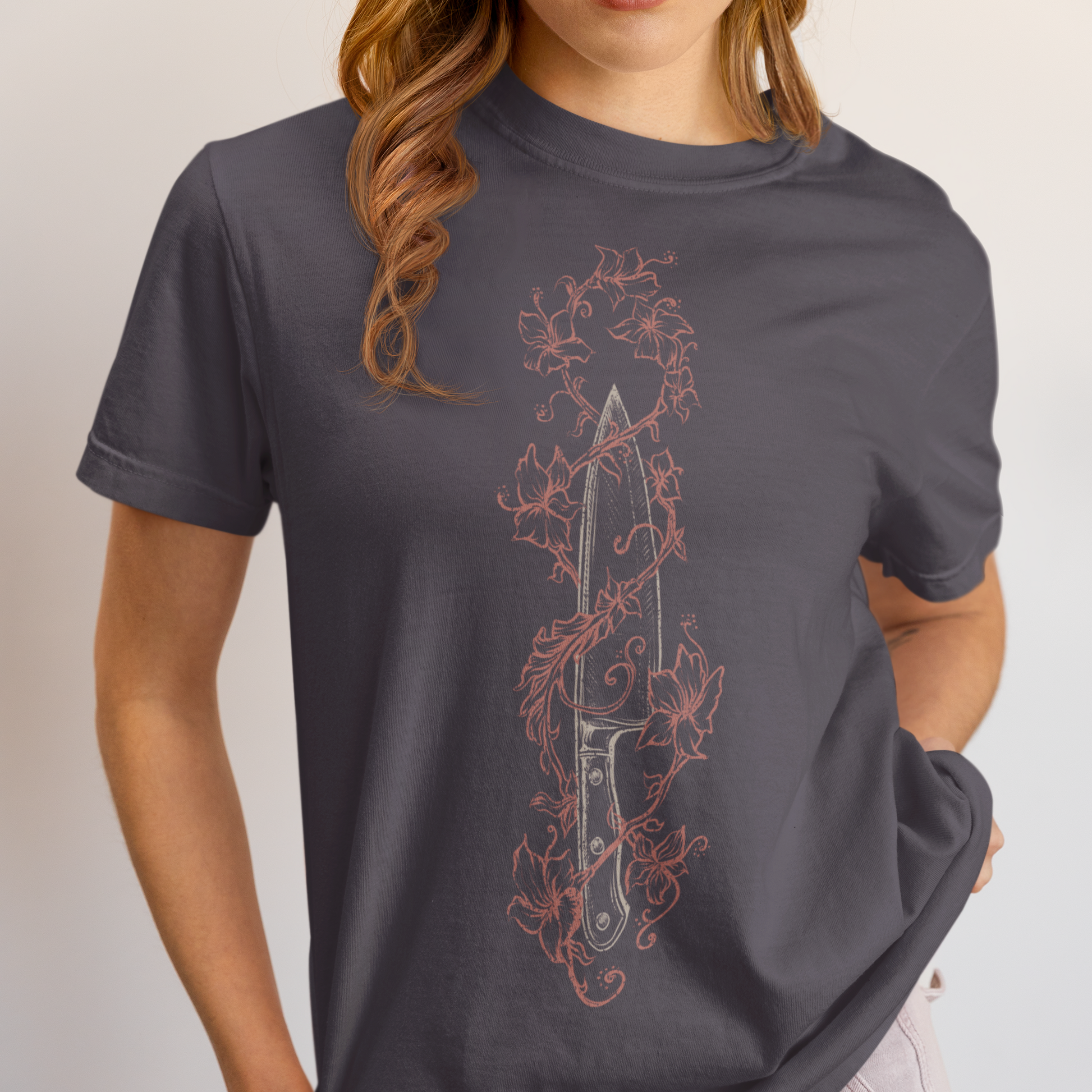 A woman wearing a dark gray t-shirt with a vertical graphic of a knife surrounded by red and pink floral vines, paired with light gray jeans against a plain light gray background.