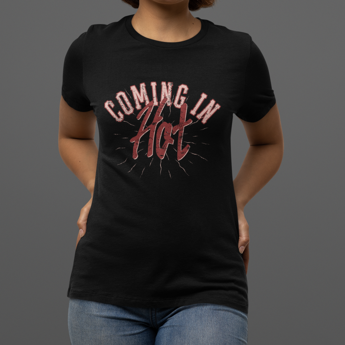 A woman wearing a black short-sleeve graphic T-shirt with the words 'COMING IN Hot' printed in pink and red across the chest, paired with blue jeans, against a solid dark gray studio background.