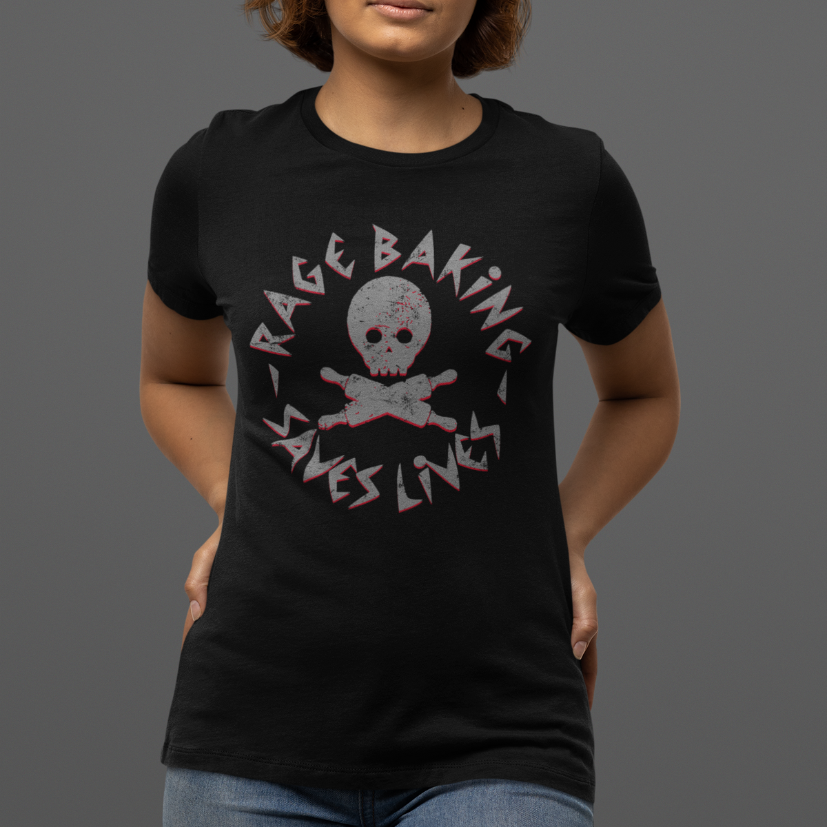 A woman wearing a black short-sleeve graphic t-shirt with a distressed pink and white 'Rage Baking Saves Lives' skull and crossbones design, paired with blue jeans, against a solid dark gray background.
