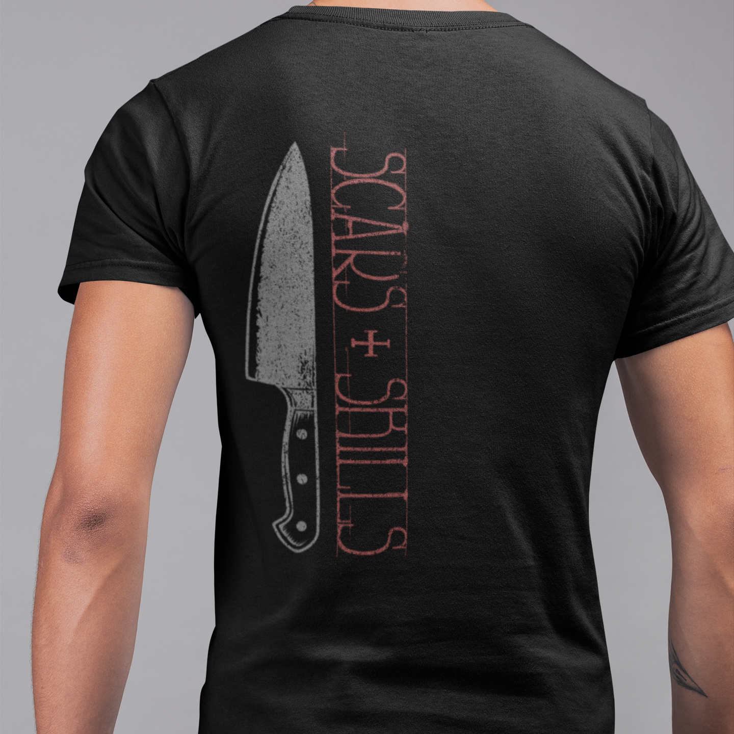 A man viewed from the back wearing a black t-shirt with a distressed graphic of a knife and the text 'SCARS + SKILLS' in red and gray.
