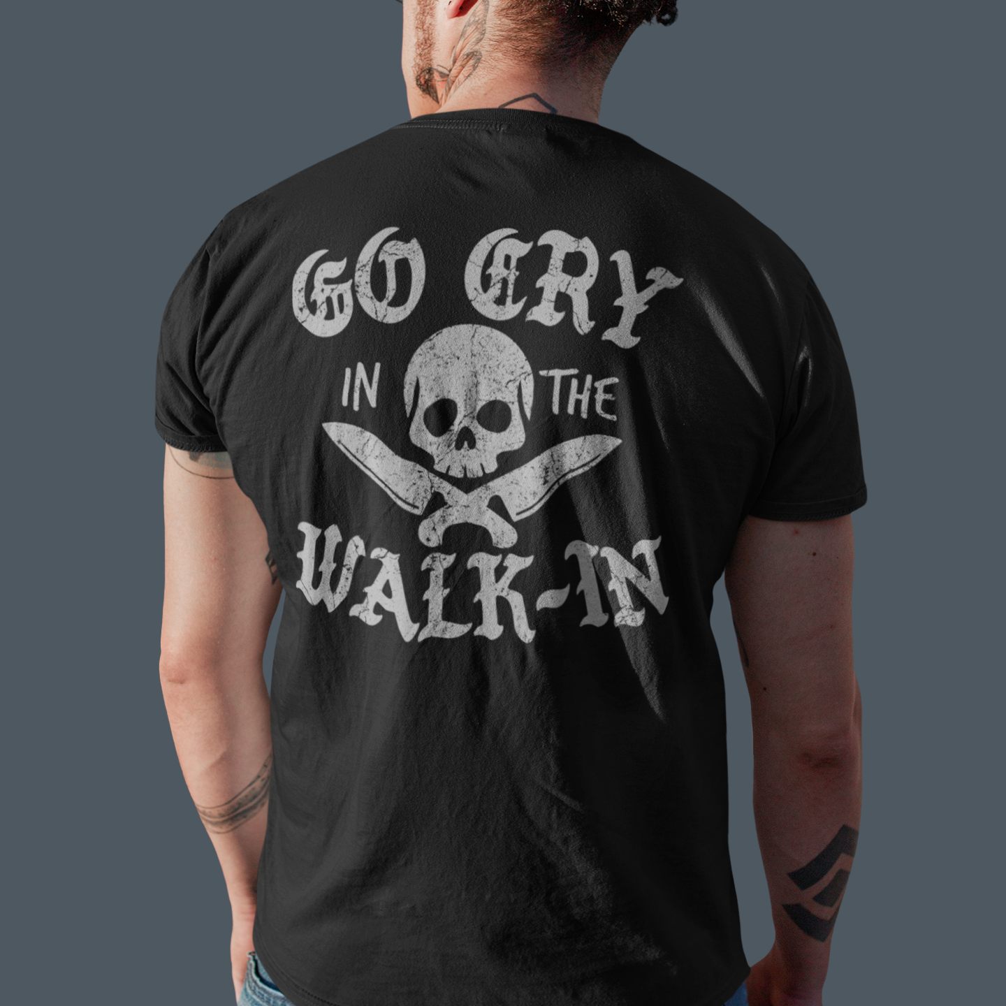 Person wearing a black t-shirt with a skull and crossbones design and text on the back, standing against a gray background.
