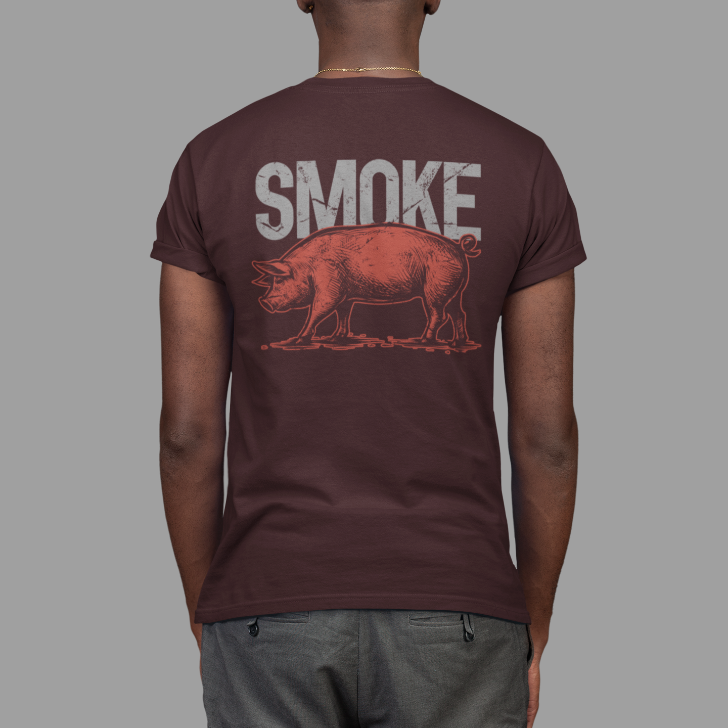 A man viewed from behind wearing a dark maroon t-shirt with a graphic print of a smoking pig and the word 'SMOKE' in distressed white letters, paired with gray pants against a solid gray background.