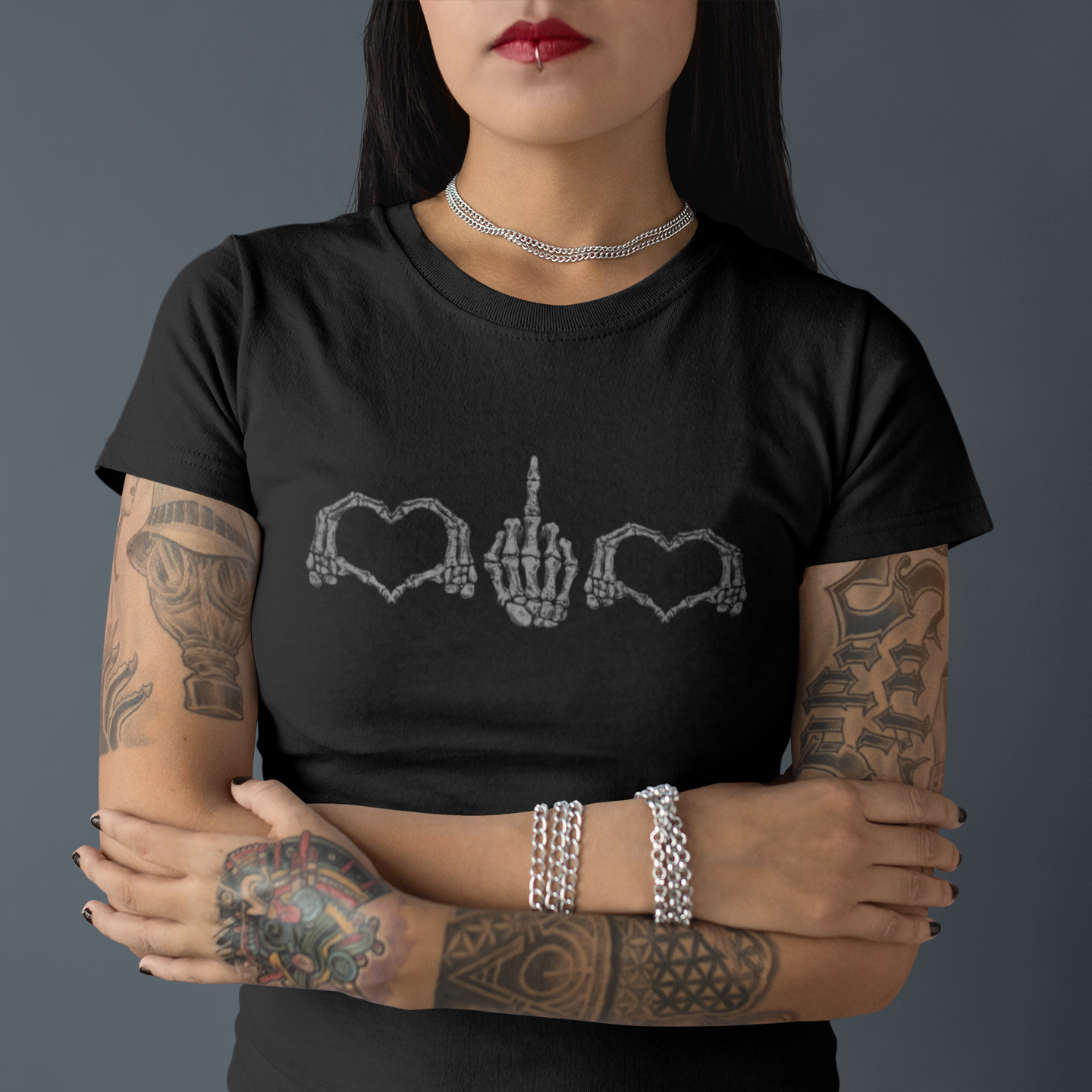A woman with tattooed arms wearing a black t-shirt with a silver graphic of skeletal hands forming hearts and a middle finger, posed against a dark gray background.