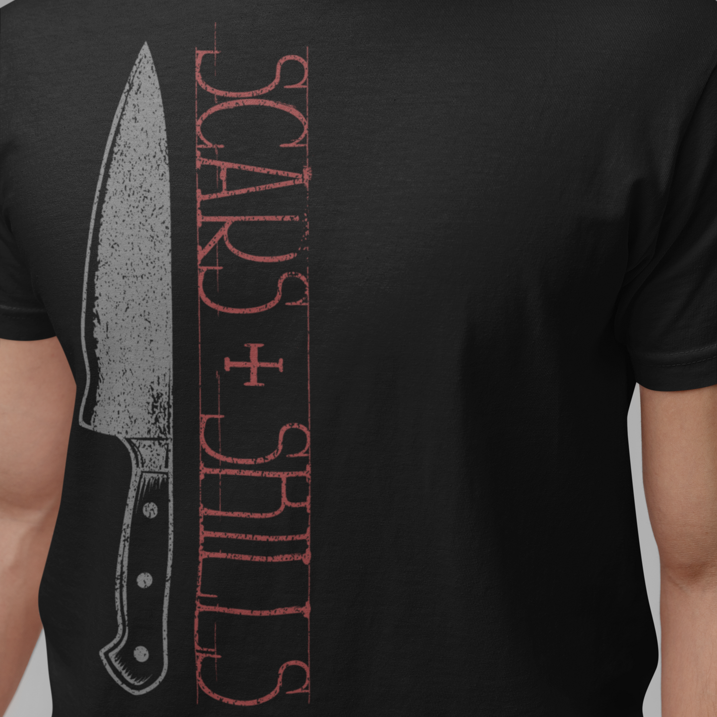 A person wearing a black short-sleeve T-shirt with a distressed graphic print featuring a knife illustration and vertical red text reading 'SCARS + SHILLS' on a plain gray background.