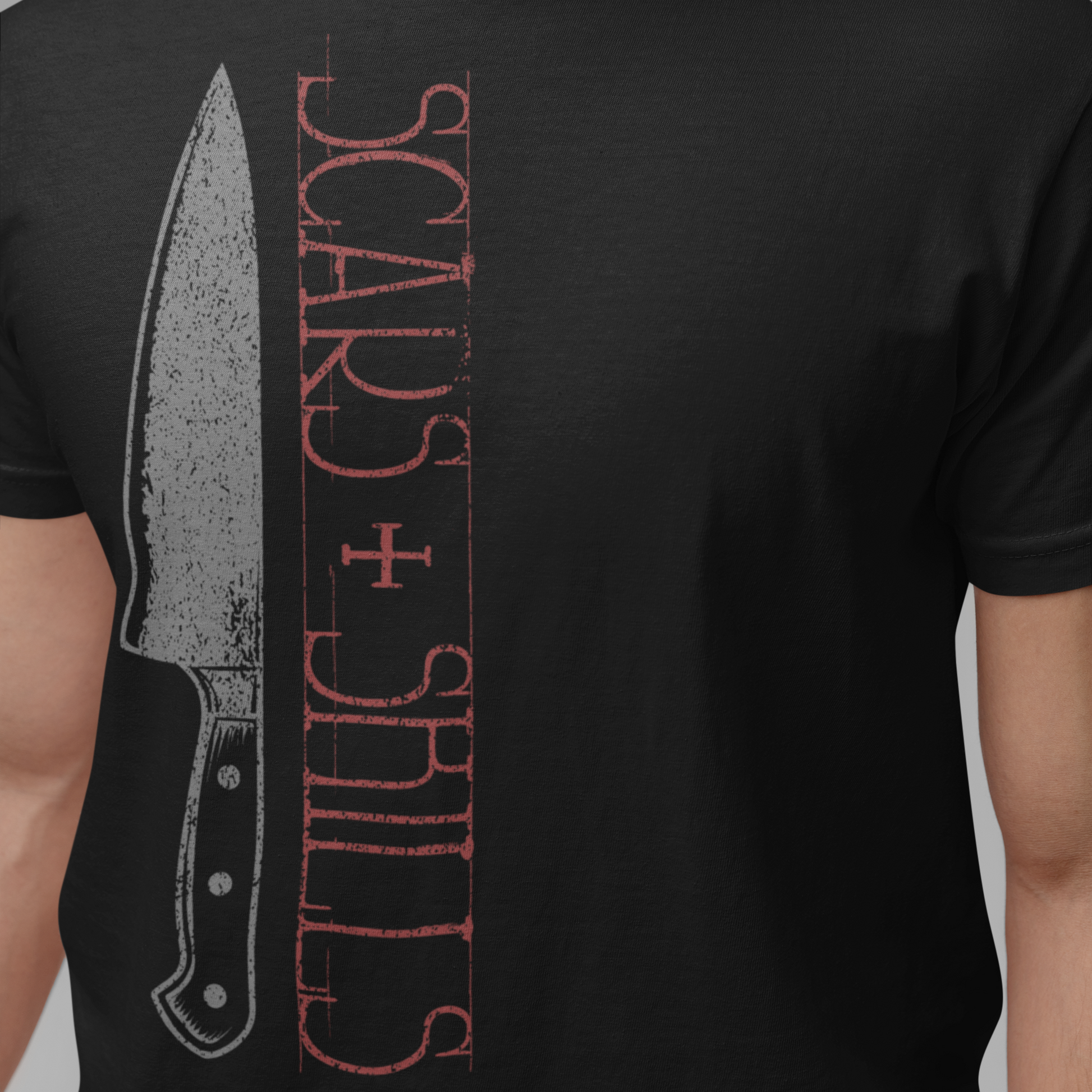 A person wearing a black short-sleeve T-shirt with a distressed graphic print featuring a knife illustration and vertical red text reading 'SCARS + SHILLS' on a plain gray background.