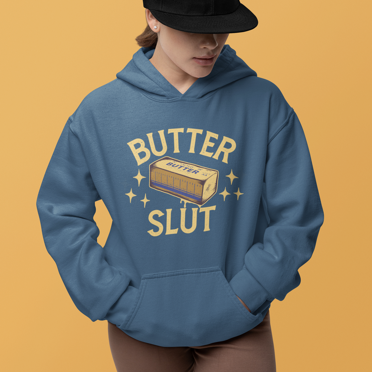 A model wearing a slate blue hoodie with a retro-style 'BUTTER SLUT' graphic on the front, paired with a black baseball cap and brown shorts, set against a solid mustard yellow background.