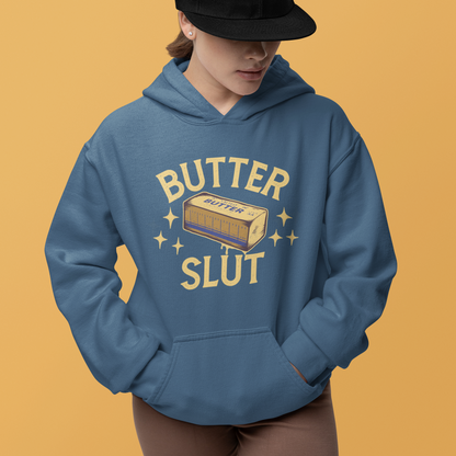 A model wearing a slate blue hoodie with a retro-style 'BUTTER SLUT' graphic on the front, paired with a black baseball cap and brown shorts, set against a solid mustard yellow background.