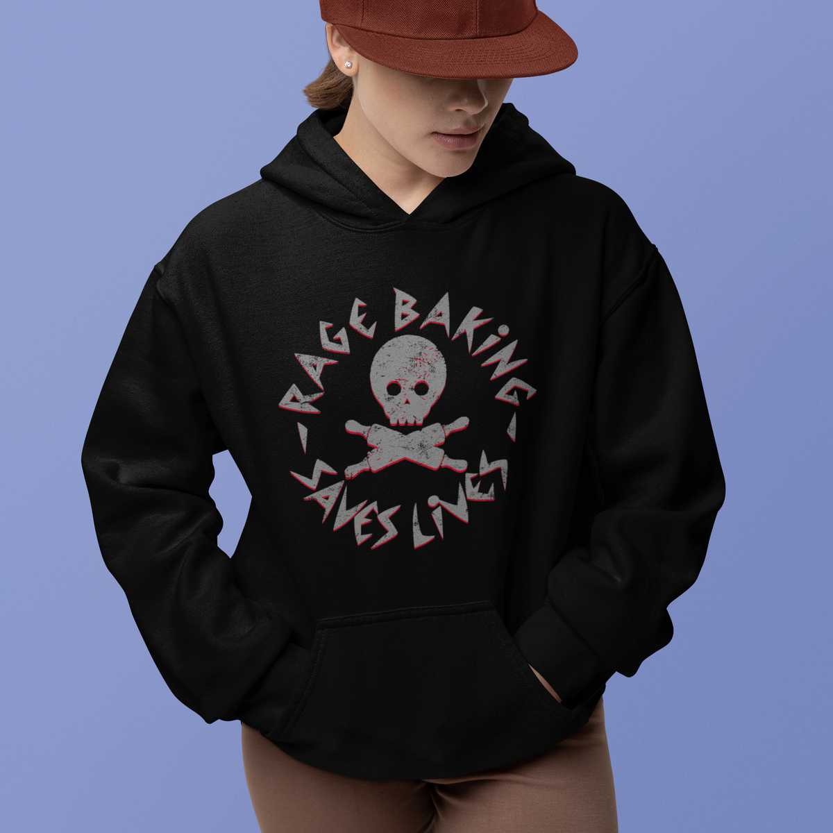 A model wearing a black hoodie with a distressed graphic that reads 'RAGE BAKING SAVES LIVES' around a skull and crossbones, paired with a rust-colored baseball cap and brown shorts, set against a solid periwinkle blue background.