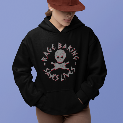 A model wearing a black hoodie with a distressed graphic that reads 'RAGE BAKING SAVES LIVES' around a skull and crossbones, paired with a rust-colored baseball cap and brown shorts, set against a solid periwinkle blue background.