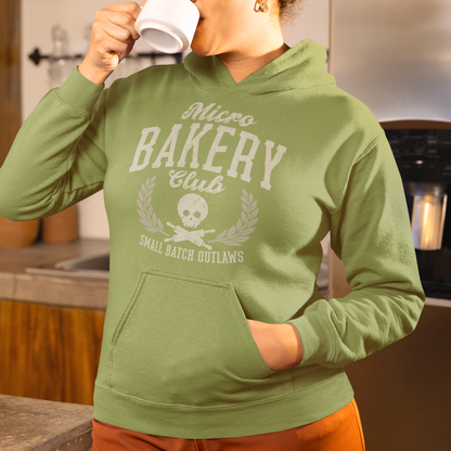A woman in a kitchen wearing an olive green hoodie with a distressed white graphic that reads 'Micro Bakery Club' and 'Small Batch Outlaws' with a skull and leaf design. She is holding a white mug to her lips.
